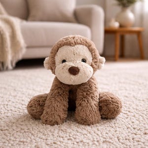 Puede incluir: Un peluche de mono con pelaje marrón y cara, orejas y vientre color crema. El animal de peluche tiene una nariz marrón en forma de corazón y está sentado sobre una alfombra de color claro.