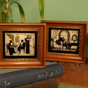 May include: Two framed silhouette art pieces with brown wooden frames. The art depicts scenes of people in a room. The frames are resting on a stack of books. The background is a green wall.