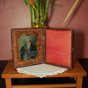 May include: An antique photograph in an open, ornate frame. The frame is dark with gold accents and a red interior. The photograph is of a man in a suit. The frame sits on a small wooden table with a white doily.