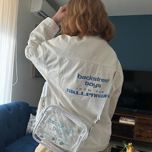 May include: A white denim jacket with blue text reading "backstreet boys" and "into the millennium". A clear crossbody bag with matching text is also visible. The jacket has a classic design with visible stitching.