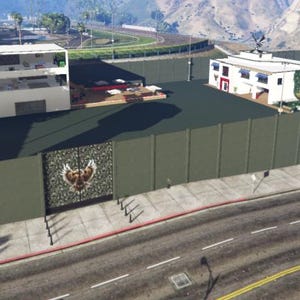 Può includere: Vista aerea di una grande proprietà con una recinzione alta verde oliva. La proprietà comprende diversi edifici, una piscina e due torri di guardia. La recinzione presenta pannelli decorativi con un disegno a mosaico. La scena è ambientata sullo sfondo di montagne e cielo sereno.