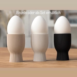 Könnte beinhalten: Drei Eierbecher, jeweils mit einem weißen Ei. Die Eierbecher sind beige, cremeweiß und schwarz. Die Becher haben ein geripptes Design. Der Text oben lautet "Einzeln oder als Set erhältlich".
