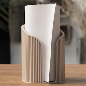 Könnte beinhalten: Ein beigefarbener Papiertuchhalter mit vertikalem Lamellendesign. Der Halter hat eine gebogene Öffnung und hält eine Rolle weißer Papiertücher. Der Halter ist aus Holz und steht auf einer Holzoberfläche.