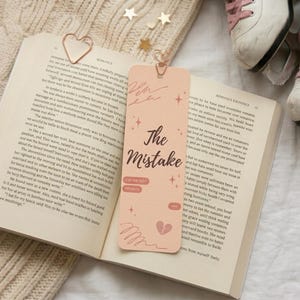 May include: An open book with a peach bookmark featuring the text "The Mistake" and a broken heart graphic. A gold heart-shaped paper clip and white ice skates with pink laces are also visible. The scene is set on a cream-colored knitted surface.