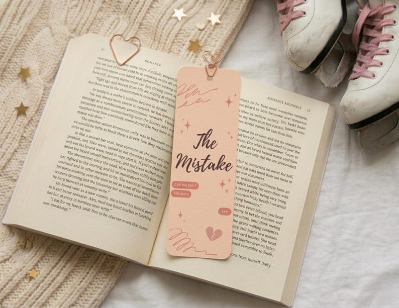 May include: An open book with a peach bookmark featuring the text "The Mistake" and a broken heart graphic. A gold heart-shaped paper clip and white ice skates with pink laces are also visible. The scene is set on a cream-colored knitted surface.