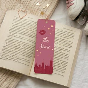 May include: A pink bookmark with the text "The Score", a lip graphic, and stars, set against a cityscape silhouette. The bookmark rests on an open book with a heart-shaped paper clip. White ice skates with pink laces are visible.