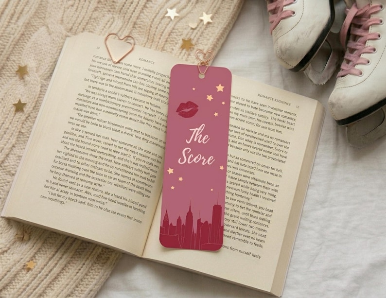 May include: A pink bookmark with the text "The Score", a lip graphic, and stars, set against a cityscape silhouette. The bookmark rests on an open book with a heart-shaped paper clip. White ice skates with pink laces are visible.