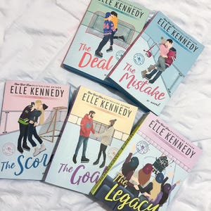 May include: Five books by Elle Kennedy are displayed. The book covers feature illustrations of couples and the titles "The Deal", "The Mistake", "The Score", "The Goal", and "The Legacy". The books are part of the "Off-Campus" series. The books are on a white surface.