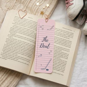 May include: A pink bookmark with the text "The Deal" and doodles of hearts, a pencil, a musical note, and a hockey stick. The bookmark is in an open book with a heart-shaped paper clip. Ice skates are in the background.