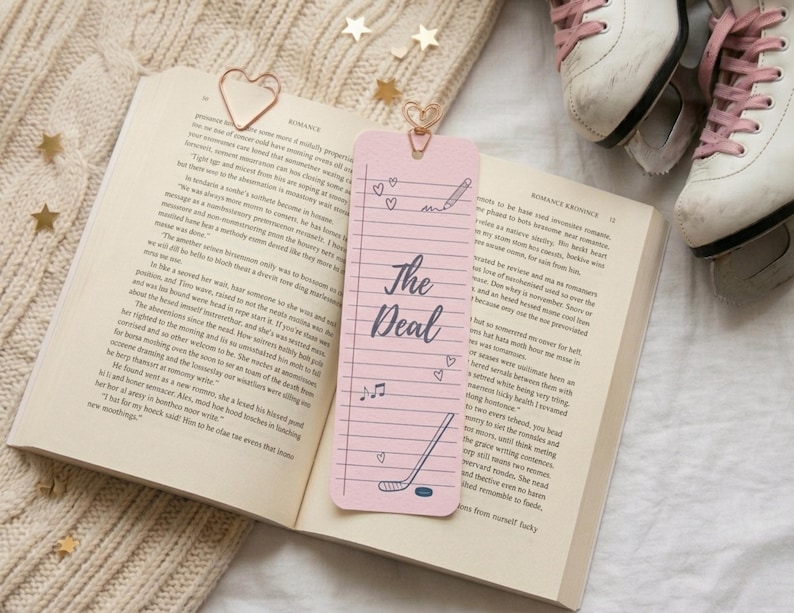 May include: A pink bookmark with the text "The Deal" and doodles of hearts, a pencil, a musical note, and a hockey stick. The bookmark is in an open book with a heart-shaped paper clip. Ice skates are in the background.