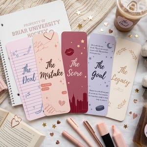 May include: A selection of romantic-themed bookmarks with phrases such as "The Deal" and "The Mistake." The bookmarks are in shades of pink, burgundy, and lavender, alongside a notebook, pens, and ice skates.