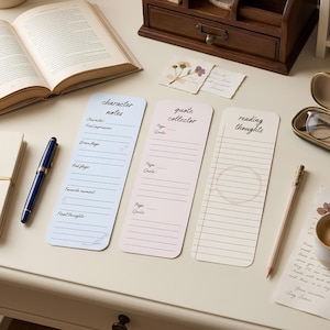 May include: Three rectangular paper bookmarks with rounded corners. The light blue one is titled character notes, the pink one is titled quote collector, and the cream one is titled reading thoughts. Each bookmark features printed lines and text.