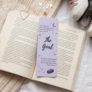 May include: A lilac bookmark with the words "The Goal" and a quote, placed in an open book. The bookmark has a hockey puck illustration. A rose gold heart paperclip and ice skates are also in view.