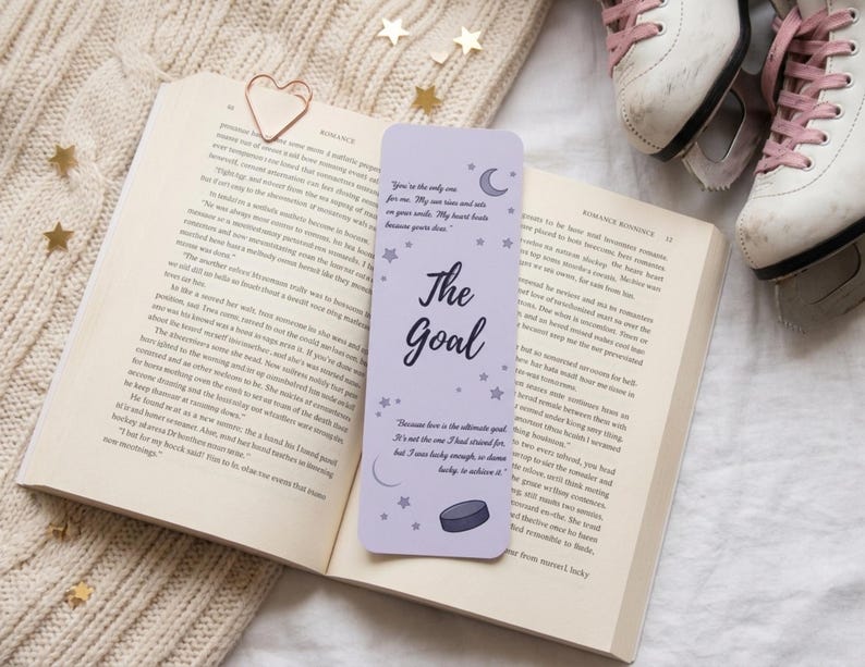May include: A lilac bookmark with the words "The Goal" and a quote, placed in an open book. The bookmark has a hockey puck illustration. A rose gold heart paperclip and ice skates are also in view.