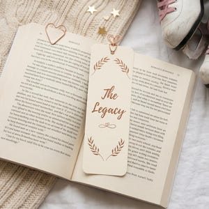 May include: An open book with a bookmark reading "The Legacy" and a heart-shaped paper clip. White ice skates with pink laces are in the background. The book rests on a cream-coloured knitted blanket with gold star decorations.