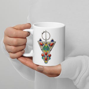 May include: White ceramic mug featuring a colorful, ornate pendant design. The pendant has red, blue, yellow, and green accents, with a silver-colored border. The mug is held by a person.