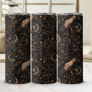 May include: A set of three stainless steel tumblers with a dark, gothic-inspired design. The pattern features hands, moons, roses, and eyes in shades of black and gold. Each tumbler has a silver straw.
