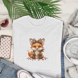 May include: A white t-shirt featuring a cute cartoon fox with a leopard print bow and scarf, holding a coffee drink. The shirt is surrounded by accessories, including sunglasses, a phone, and a pair of headphones.
