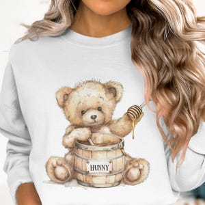May include: A watercolor illustration of a teddy bear with a honey dipper and a barrel of honey. The bear is light brown and sitting in front of a wooden barrel with the word "HUNNY" on it. The honey is dripping from the dipper.