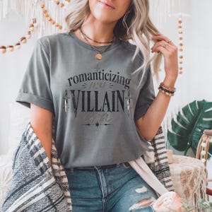 Grey t-shirt with "Romanticizing my Villain Era" text and sword graphics.