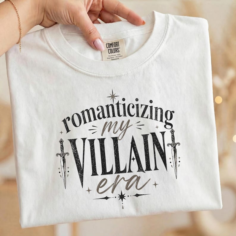 White t-shirt with "romanticizing my villain era" text and sword graphics.