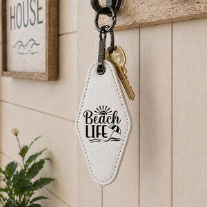 May include: A white, diamond-shaped keychain with the words "Beach Life" printed in black, hanging from a black hook. The keychain has a stitched border and a small umbrella graphic. A set of keys is attached.