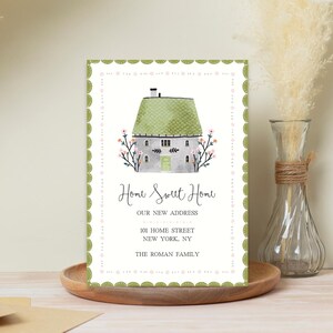 May include: A watercolour new home announcement card. The card features a house with a green roof and pink flowers. The text reads "Home Sweet Home" and includes the new address and family name. The card has a green and pink border.