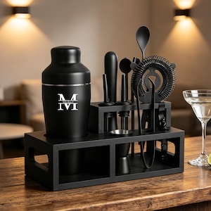 Puede incluir: Un juego de herramientas de bar negro con una coctelera, un medidor y un colador, exhibido en una barra de madera. La coctelera tiene una banda plateada y un monograma blanco. Una copa de martini con una rodaja de lima y dos limas están en la barra.