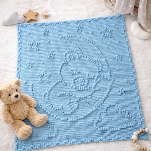 May include: A light blue square crochet blanket featuring a textured bobble stitch design of a bear sitting on a crescent moon surrounded by stars and clouds. A tan plush teddy bear sits on the bottom left corner of the blue blanket.