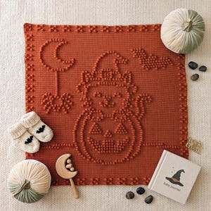May include: A square, burnt-orange crochet blanket featuring a textured bobble-stitch design of a cat wearing a witch hat, a crescent moon, a spider, and a bat. The blanket is surrounded by two velvet pumpkins, cream-colored baby booties, a wooden moon rattle, and a baby journal.