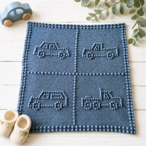Peut inclure: Une couverture pour bébé en crochet bleu avec quatre motifs de voitures. La couverture a une bordure texturée. Un jouet de voiture en bois et une paire de chaussons crème sont également visibles.