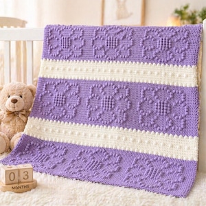 May include: A handmade crochet baby blanket in lavender and cream. The blanket features a repeating floral pattern in lavender, with cream-coloured stripes. The blanket is displayed on a white cot, with a teddy bear and a wooden block calendar that reads "03 months."