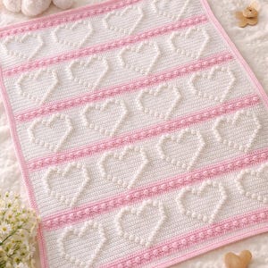 May include: A white and pink crocheted baby blanket with a heart design. The blanket has rows of white hearts separated by pink stripes and a pink border. A stuffed bunny, baby shoes, and a rattle are also visible in the background.