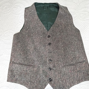 May include: A brown and grey tweed vest featuring a speckled pattern, a V-neckline, and five dark buttons down the center. The garment includes two front welt pockets and is lined with a dark green fabric that has a subtle repeating logo pattern.