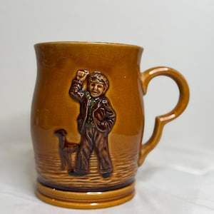 Puede incluir: Taza de cerámica marrón con asa, con un relieve en relieve de un niño con un perro. La taza tiene un acabado brillante y una base ancha. El niño lleva sombrero y chaqueta. Un objeto de colección de estilo vintage.