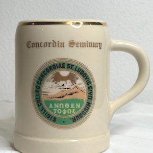 May include: Cream-colored ceramic stein with a gold rim and handle. The stein features the text "Concordia Seminary" and a circular emblem with text and a landscape scene. The emblem reads "COLLEG. CONCORDIAE. ST. LUDOVIC. CIVIT. MISSOUR. 1915".
