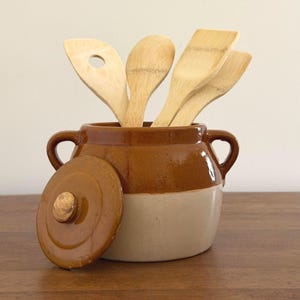 May include: A two-toned ceramic crock with a brown glazed upper section and a cream-colored base, featuring two side handles and a matching removable lid. Several light-colored wooden kitchen utensils are placed inside the container.