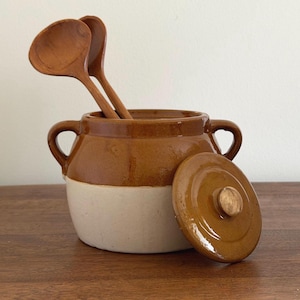 May include: A two-toned ceramic pot with a brown glazed upper section and a cream-colored base. The pot features two side handles and a matching brown lid with a central knob. Two wooden ladles are placed inside the pot, extending upwards.