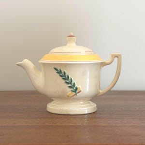 May include: Cream-colored ceramic teapot with a lid and handle. The teapot features a yellow band around the lid and a green and yellow floral design on the front. The teapot is on a pedestal base.