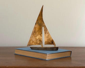May include: A metallic bronze-toned sailboat sculpture featuring a tall, pointed sail and a solid base. The sculpture has a weathered, mottled finish with dark and light brown patches, depicting a stylized vessel with a single mast and sail.