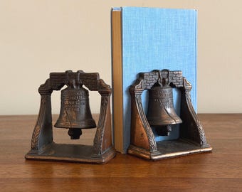 May include: A pair of bronze-toned metal bookends shaped like the Liberty Bell, each featuring a detailed bell suspended within an arched frame with brick-like textures and leaf motifs, supporting a single light blue hardcover book between them.