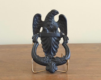 May include: A black cast iron door knocker featuring a detailed eagle perched atop a shield-shaped plate with ornate scrollwork at the base. The eagle is depicted with wings partially spread and head turned to the right, showcasing a textured finish.