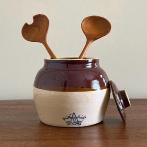 May include: A two-toned stoneware crock with a dark brown glazed upper section and a cream-colored lower base. A blue stamped crown logo with the number 2 is visible on the front. Two wooden spoons are inside, and a matching lid rests beside it.
