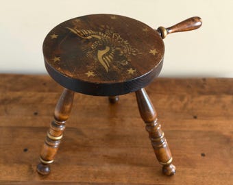 May include: A dark brown wooden three-legged stool with a round seat featuring a gold-colored graphic of a winged emblem and stars. The stool includes a single wooden handle extending from the side of the seat and turned wooden legs.