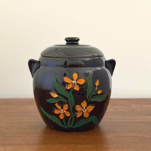 May include: A dark brown ceramic jar with a lid and two side handles. The front of the jar features a hand-painted design of orange flowers with green leaves and stems. The surface of the jar has a glossy finish and a rounded, bulbous shape.