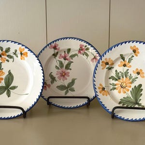 May include: Three round ceramic plates with a white base and a blue scalloped rim. Each plate features a hand-painted floral motif with green leaves and yellow or pink petals. The plates are displayed upright on individual black metal stands.