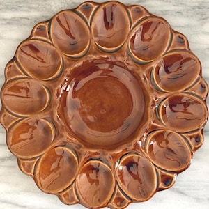 May include: A brown ceramic egg serving platter with a glossy finish. The platter has 12 egg-shaped indentations around the edge, with a circular indentation in the center. The platter is approximately 12 inches in diameter.