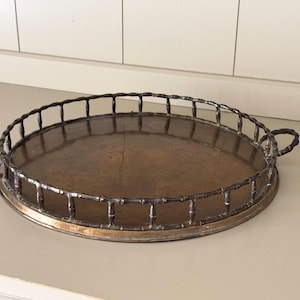 May include: A round metal serving tray with a dark, weathered bronze finish. The tray features a raised gallery rail designed to resemble bamboo stalks and includes two matching circular handles on opposite sides for easy carrying.