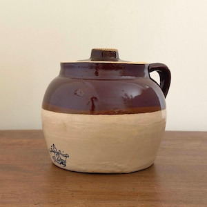 May include: A two-toned stoneware crock with a dark brown glazed upper section and a light beige lower section. The vessel features a single side handle, a matching lid, and a blue stamped mark on the lower front displaying the number two.
