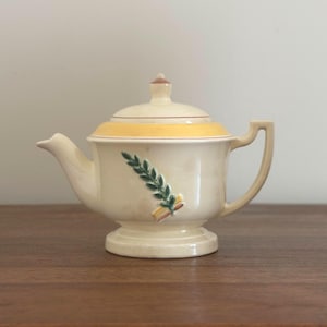 May include: Cream-colored ceramic teapot with a lid and handle. The teapot features a yellow band around the middle and a green and yellow leaf design on the front. The teapot is on a pedestal base.